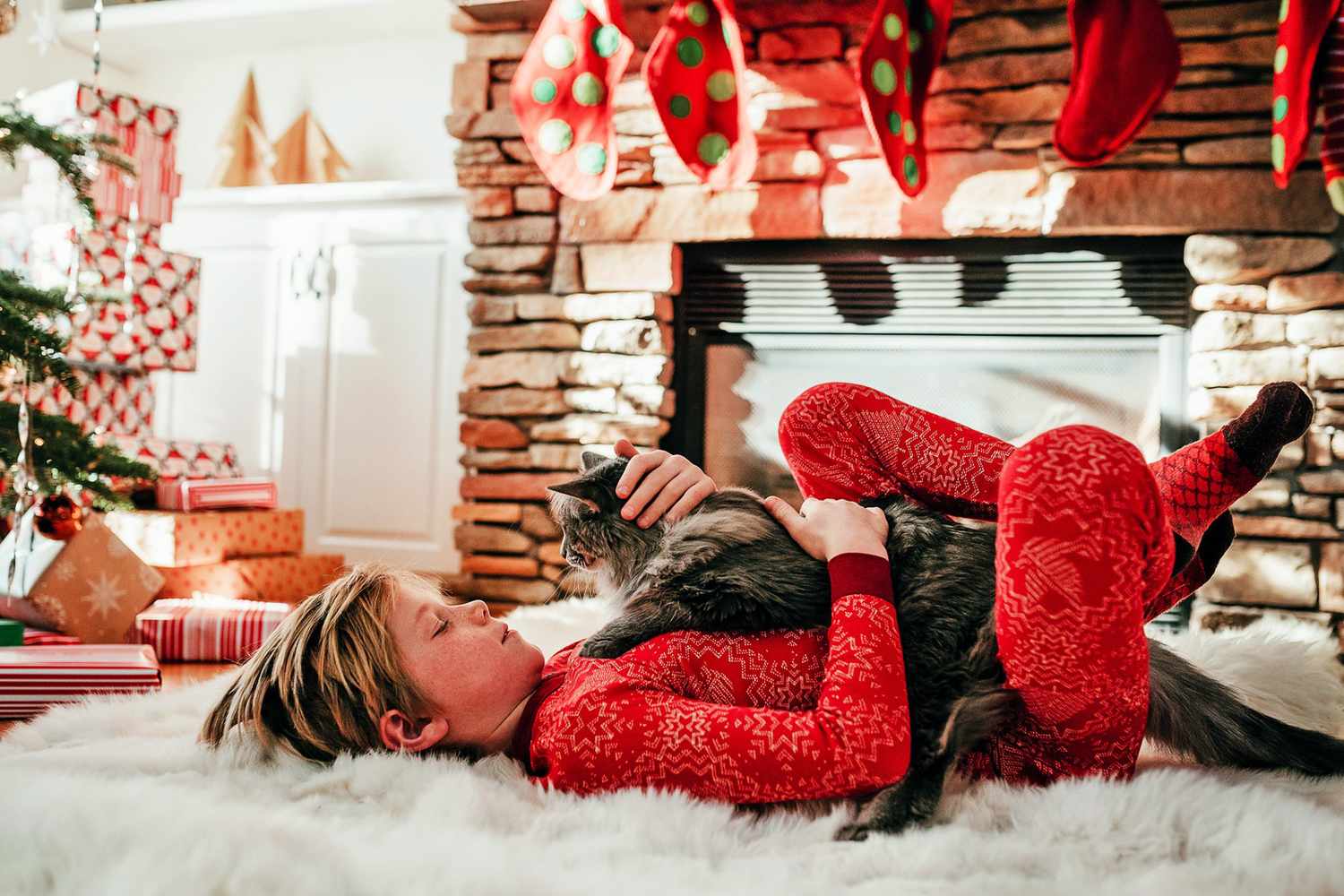 ragazzo sdraiato su un soffice tappeto bianco con un gatto sul petto accanto a un albero di Natale